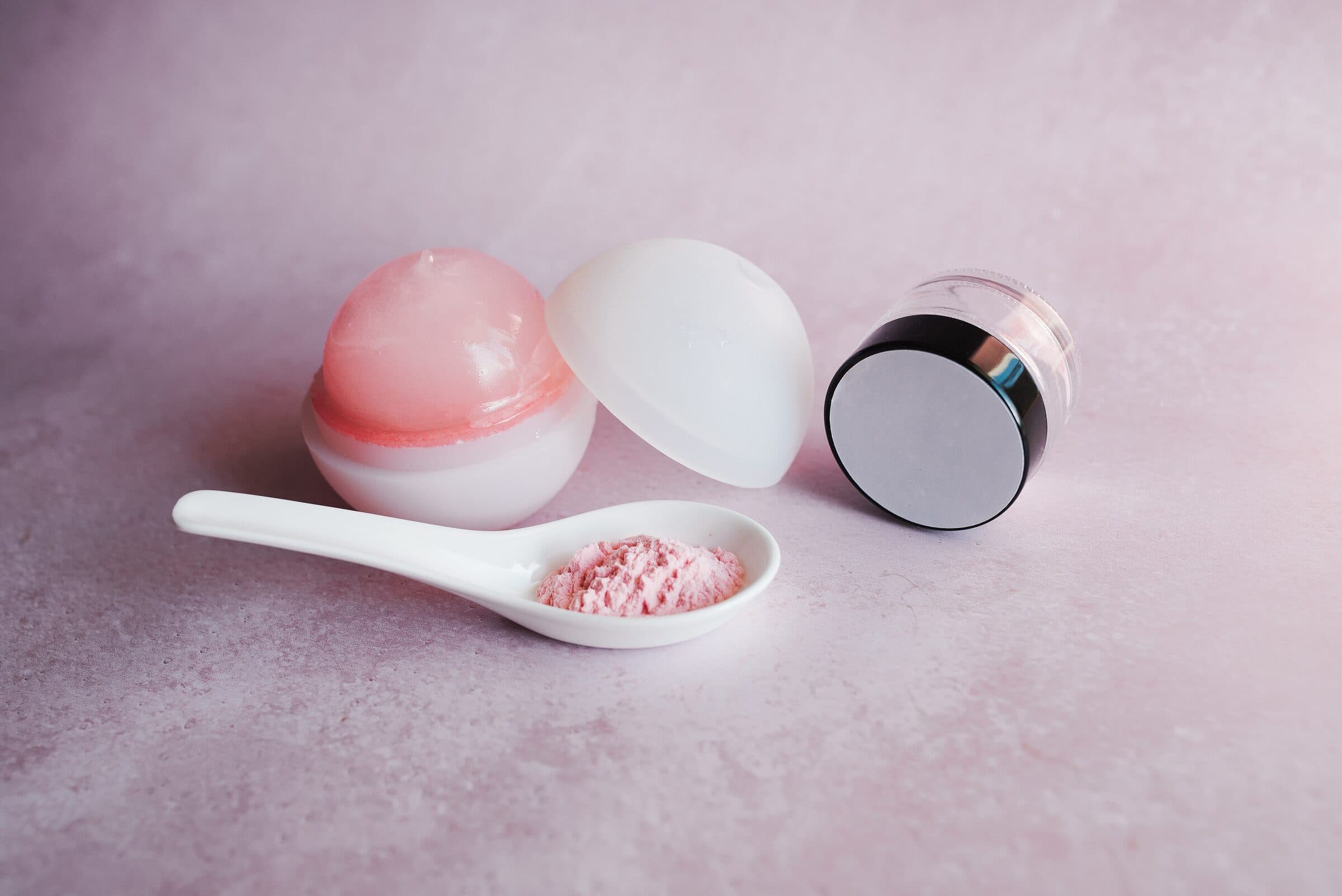 Zwei Tiegel mit Pflegeprodukten, einer offen mit rosafarbenem Balsam, daneben ein weißer Löffel mit rosa Puder auf zartrosa Grund